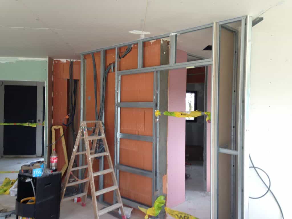 Chantier intérieur avec ossature métallique et cloisons en placo (rose, vert) et briques. Câbles électriques et échelle en bois.