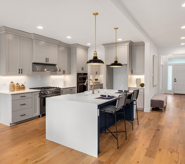 Cuisine moderne avec armoires grises, comptoirs blancs, plancher bois clair, îlot central bleu marine, et luminaires pendants dorés.