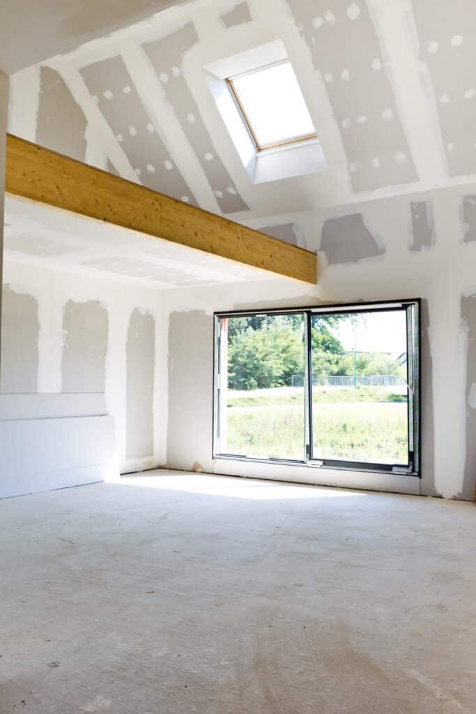 Intérieur de maison neuve en travaux. Murs et plafond en placo, poutre bois, velux et grande baie vitrée. Sol béton brut.