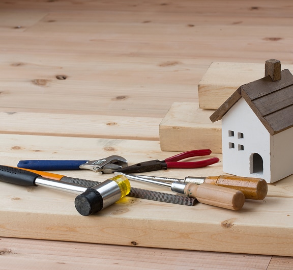 Outils de bricolage variés (clé, pinces, marteau, tournevis) et maquette de maison sur planches de bois clair.