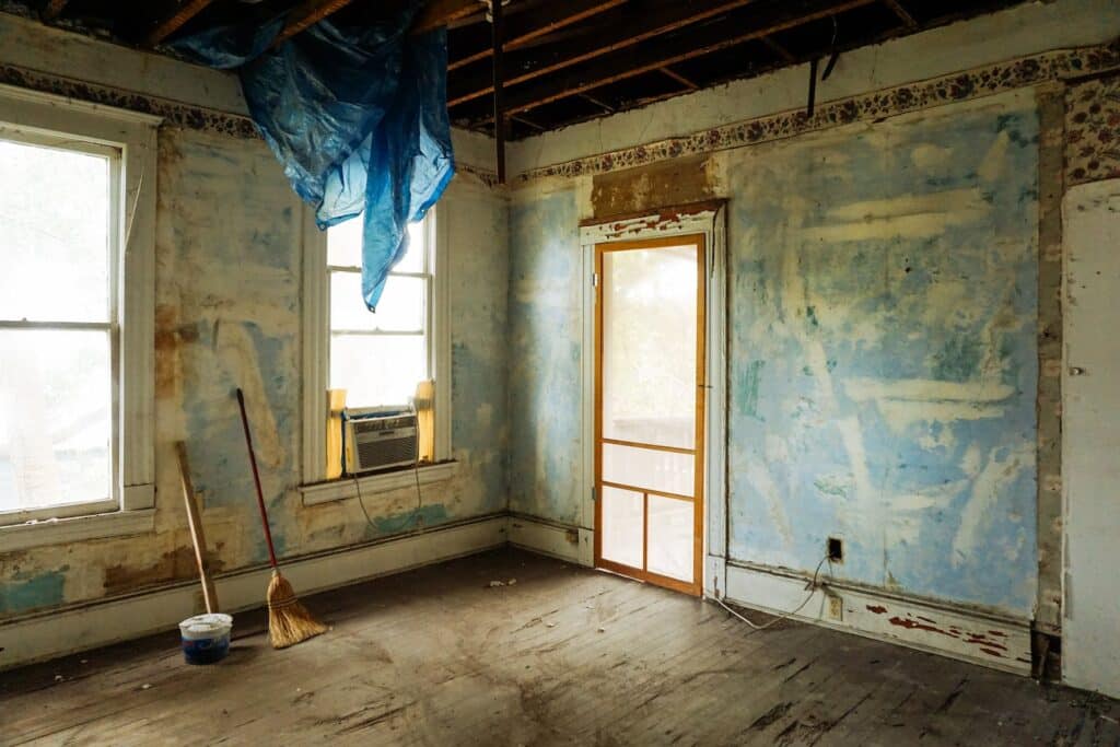 Pièce en rénovation avec murs bleus écaillés, plafond nu, sol en bois sale. Tarp bleu, balai et climatiseur sous fenêtres lumineuses.