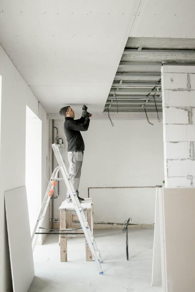 Homme installant des plaques de placo au plafond avec une visseuse, debout sur un échafaudage de fortune. Chantier de rénovation.