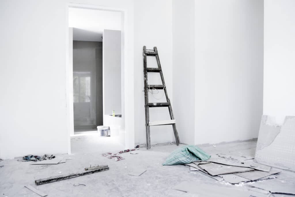 Rénovation Intérieure : Espace Inachevé et Échelle Intérieur en rénovation avec murs blancs, échelle en bois, sol en béton et débris de chantier. Une porte mène à une pièce carrelée.