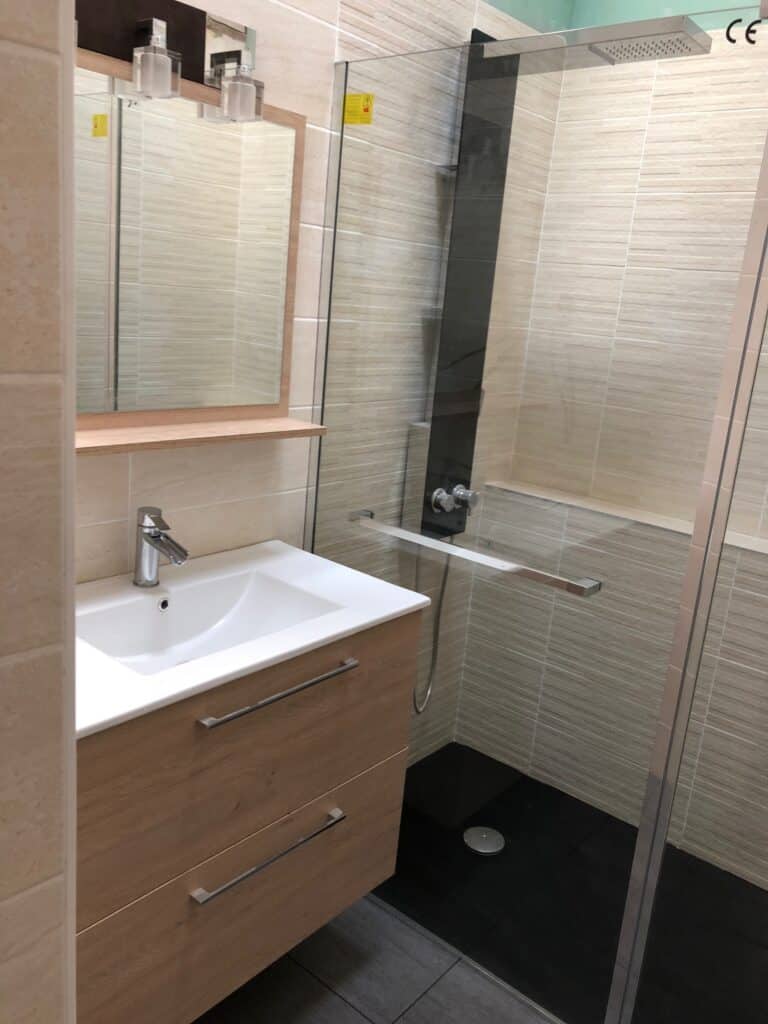 Salle de bain moderne: meuble vasque bois clair, évier blanc, miroir éclairé. Douche vitrée, carrelage beige, sol noir. Autocollant attention sur verre, marque CE.