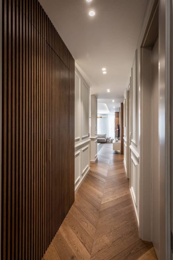 Long couloir moderne avec mur de tasseaux en bois foncé, boiseries blanches et parquet chevrons. Plafond éclairé par des spots.
