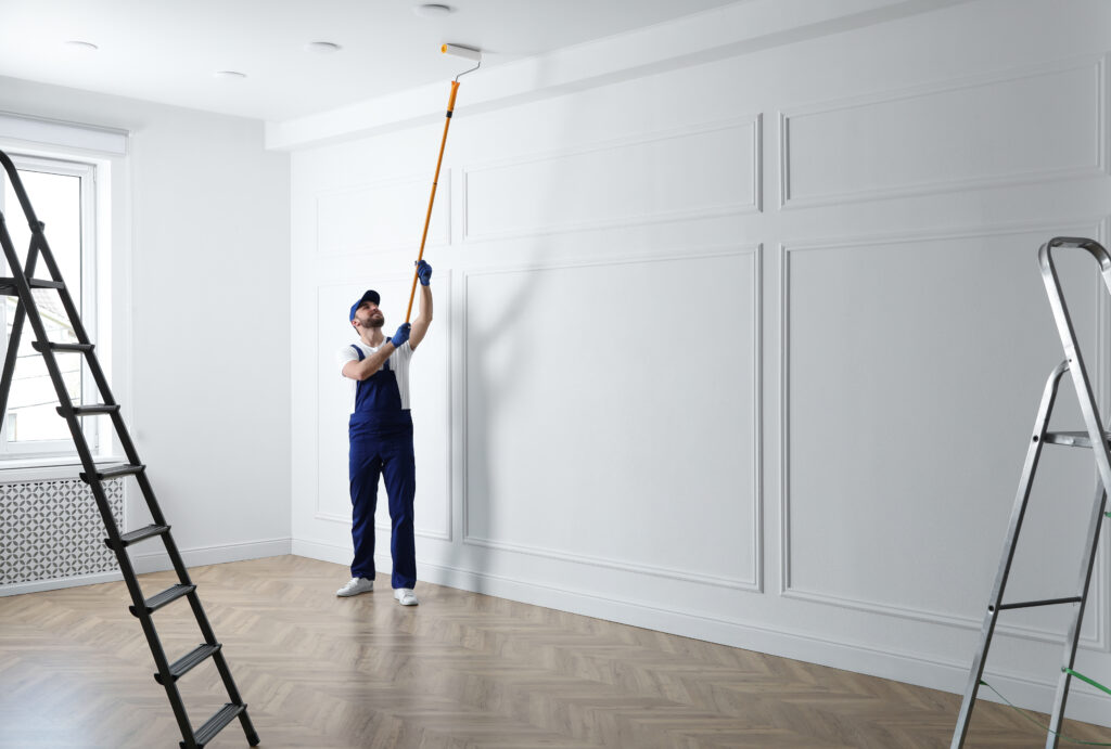 Peintre en salopette bleue appliquant de la peinture blanche au plafond d'une pièce rénovée, murs blancs moulurés, parquet chevrons, échelles.