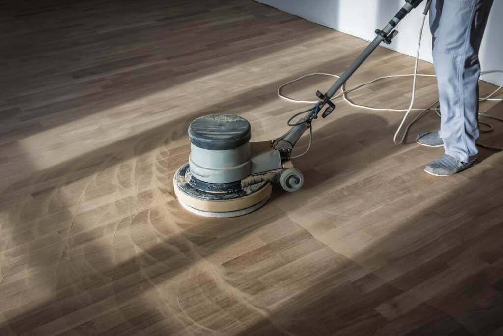 Homme ponçant un parquet en bois clair avec une machine rotative. Des traces de ponçage et de poussière sont visibles sur le sol.